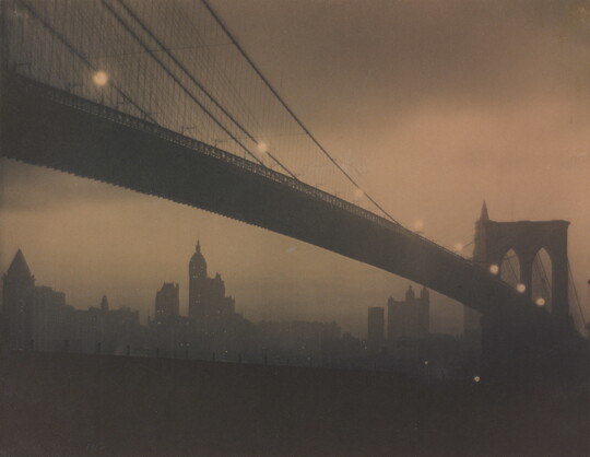 A sepia-toned photograph of a long bridge with a foggy cityscape in the background taken at night.
