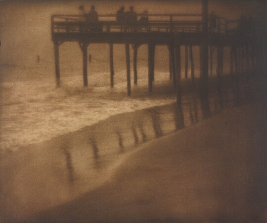 A sepia-toned photograph of people standing on a pier as waves crash beneath it.