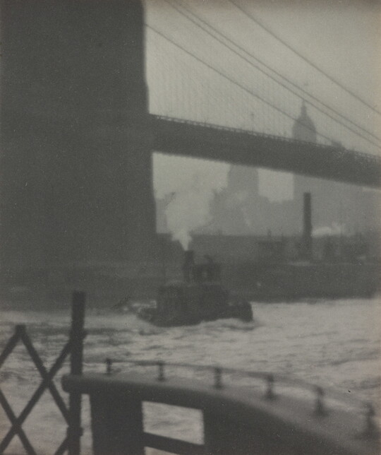 A black-and-white photograph of a suspension bridge seen from below, a tugboat on the water, and a city skyline in the distance.