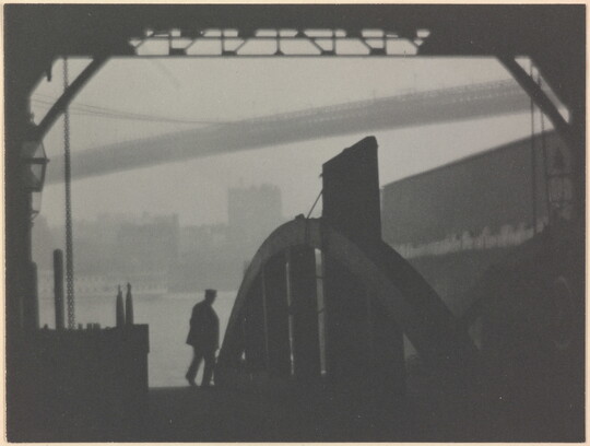 A black-and-white photograph of the silhouette of a person walking on a dock with a hazy city and a large bridge in the background.
