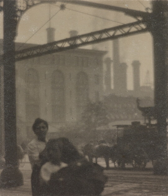 A black-and-white photograph of people and horse-drawn carriages on a street in front of a factory.