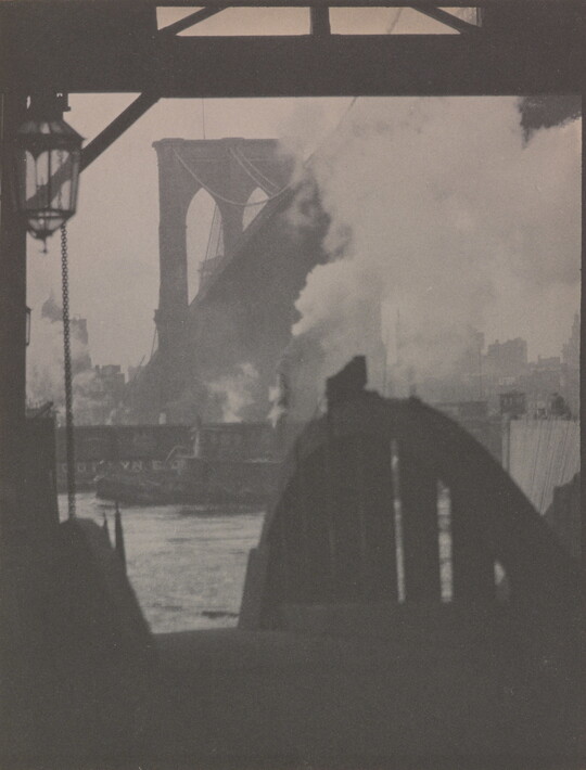 A black-and-white photograph of a dock in silhouette with a hazy city and a large bridge in the background.