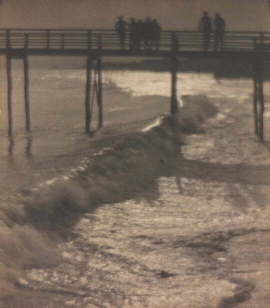 A sepia-toned photograph of people standing on a pier as waves crash beneath it.