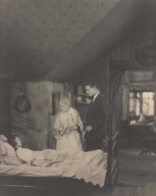 A black-and-white photograph of a White woman lying in bed as an elderly woman and a tall young man watch over her.