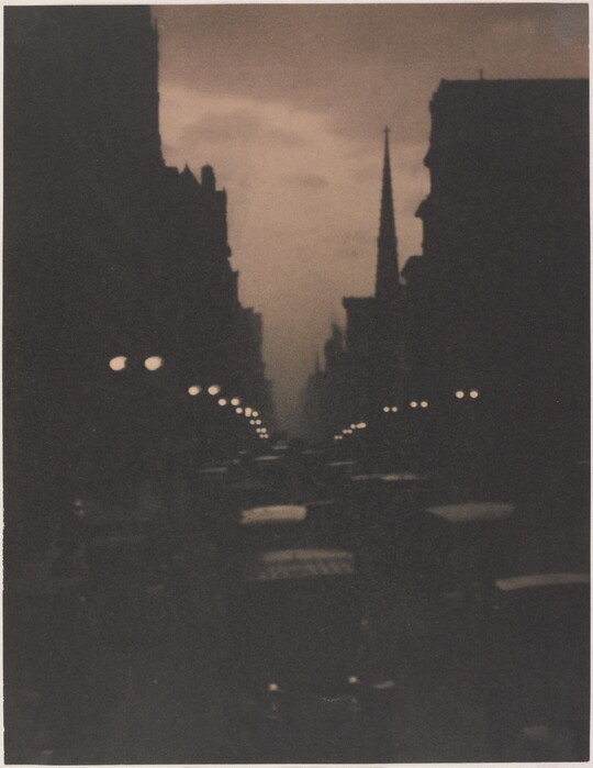 A sepia-toned photograph of a busy city street at dusk illuminated by street lamps and car lights.