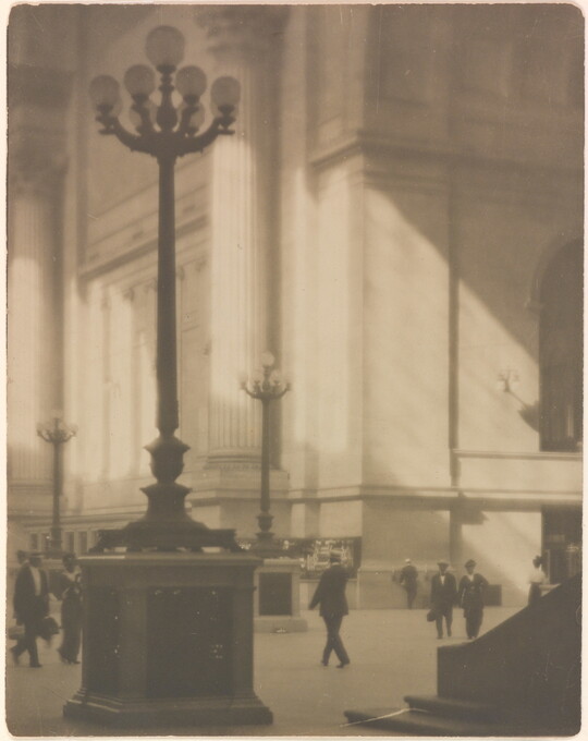 A sepia-toned photograph of people walking through a sunlit plaza with towering light posts and columns.