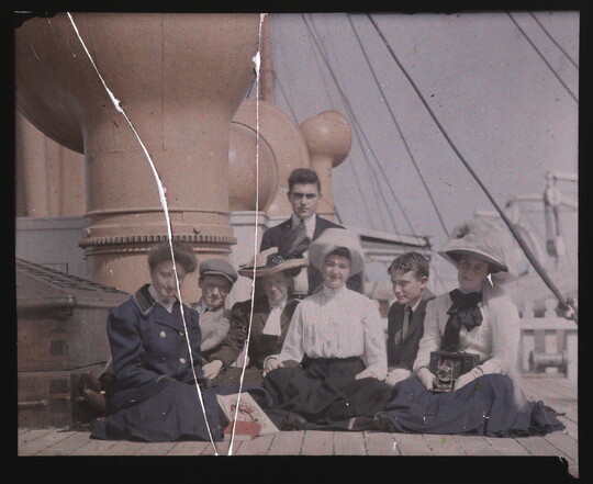 A color photograph of a group of young, fair-skinned, adults sitting on the sunny deck of a ship.