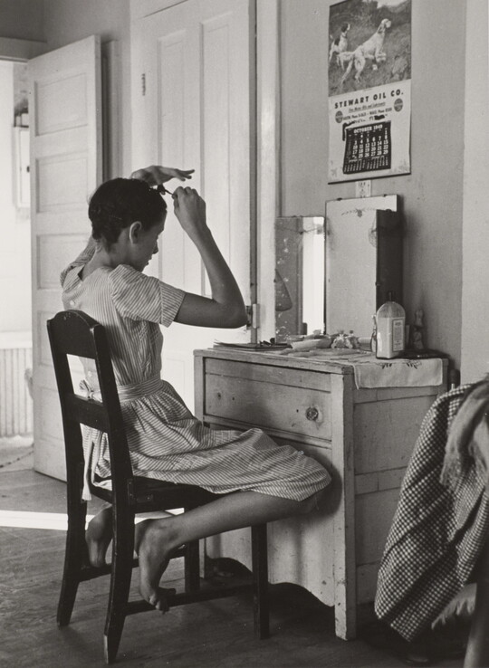 A black-and-white photograph of a White teenage girl seated in front of a mirror on top of a dresser fixing her hair.
