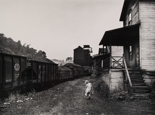 A black-and-white photograph of a young girl carrying a pail between a train and some clapboard buildings.