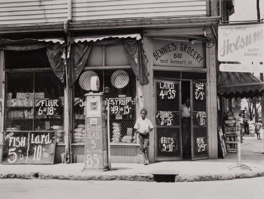 A black-and-white photograph of a dark-skinned boy standing in front of a grocery store.