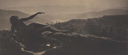 A sepia-toned photograph of a nude woman in shadow lying on a hilltop with cascading hills behind her.