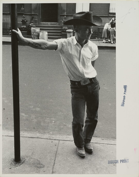 A black-and-white photograph of a White man in a cowboy hat leaning against a metal pole on a city sidewalk.