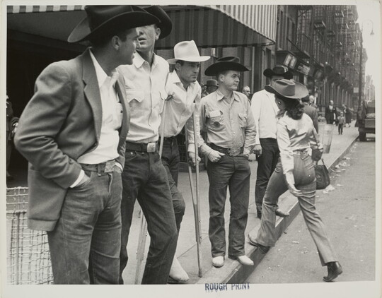 A black-and-white photograph of a group of White cowboys standing on a city curb as a woman in a cowboy hat steps up on the curb to the sidewalk.
