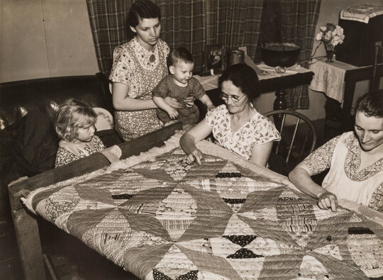 A black-and-white photograph of two seated White women quilting, a woman with a child on her lap, and another child seated next to her.