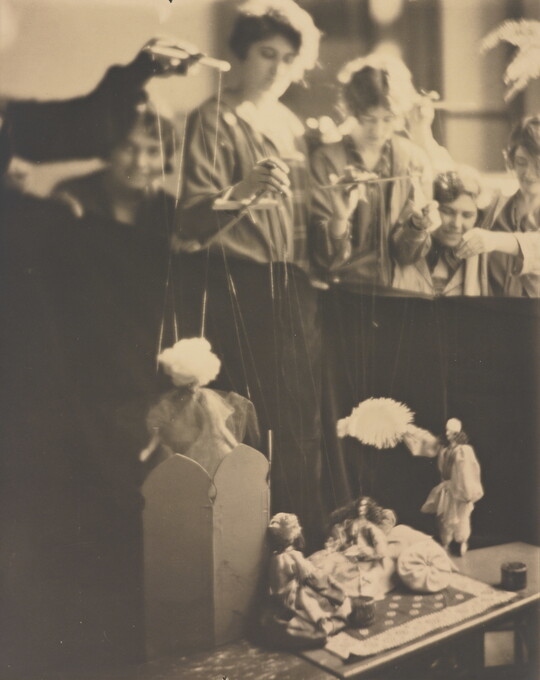 A sepia-toned photograph of several White women working marionettes above a puppet stage.