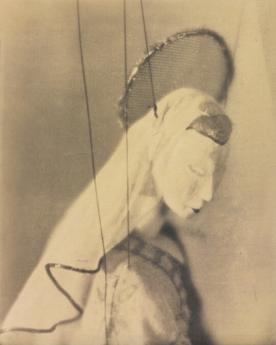 A sepia-toned close-up photograph of the head and shoulders of a marionette with a halo and veil.