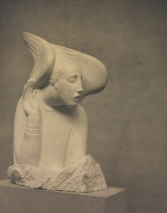 A sepia-toned photograph of a white stone sculpture of an abstracted human figure with wings around the sides of its head.