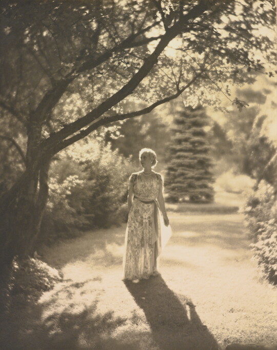 A black-and-white photograph of a light-skinned woman in a flowing dress walking among trees with the sun at her back.