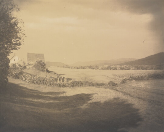 A sepia-toned photograph of an open field with a house and a barn in the distance.