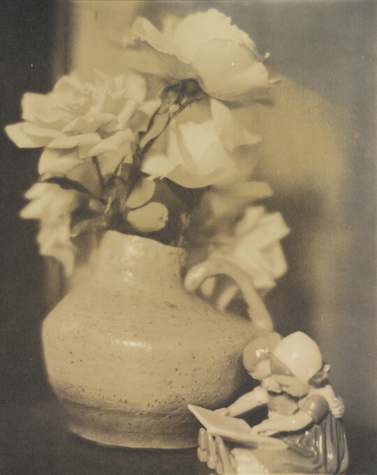 A sepia-toned photograph of roses in a vase and a figurine of two young children reading a book.