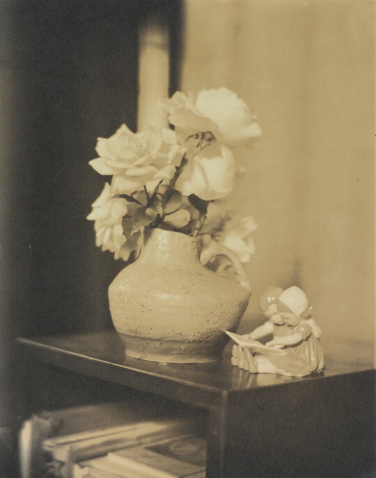 A sepia-toned photograph of roses in a vase and a figurine of two young children reading a book on a shelf.