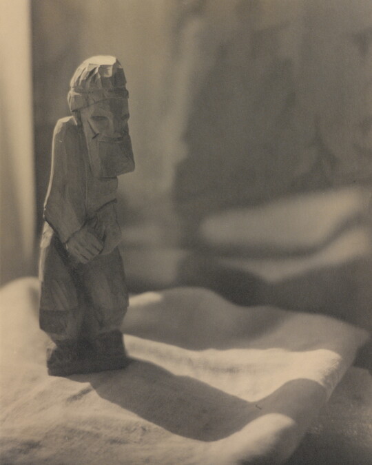 A sepia-toned photograph of a rough-hewn sculpture of a bearded man standing on a piece of folded fabric.