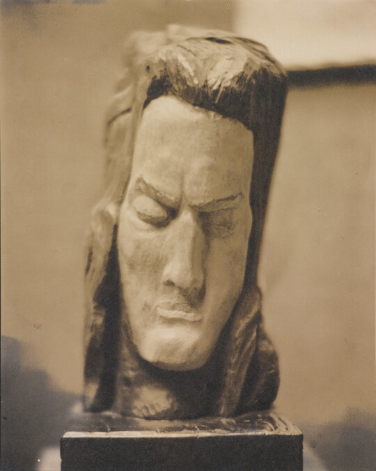 A sepia-toned photograph of a rough-hewn sculpture of a man's head resting on a pedestal.