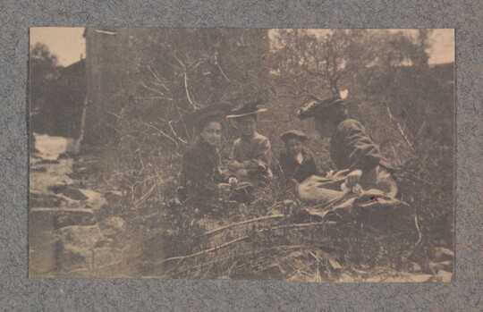 A black-and-white photograph of a group of four light-skinned women wearing hats and dresses sitting among trees and bushes.