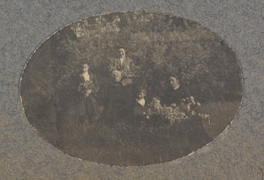 An oval-shaped black-and-white photograph of a group of light-skinned women and children holding flower bouquets in a natural setting.