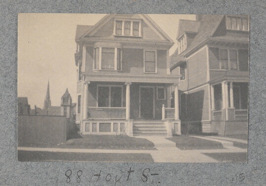 A black-and-white photograph of a three-story wood house, with a front porch and steps leading to a sidewalk in front.
