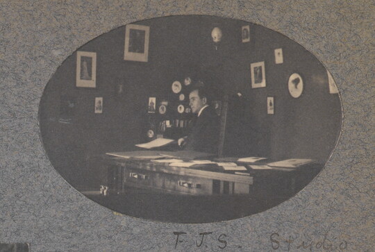 An oval-shaped black-and-white photograph of a White man seated behind a desk; framed pictures hang on the walls behind him.
