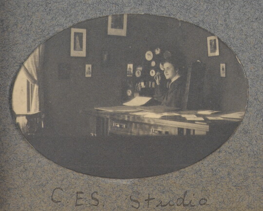 An oval-shaped black-and-white photograph of a White woman seated at a desk; framed pictures hang on the walls behind her.