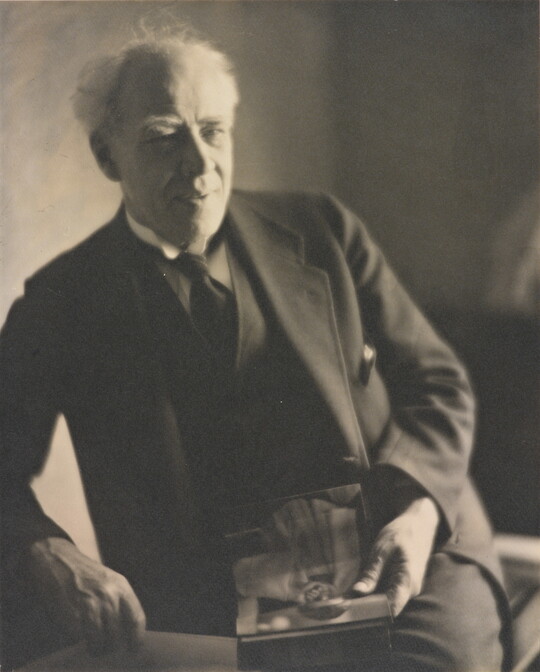 A black-and-white photograph of an older White man with white hair, wearing a dark suit and tie, leaning on one arm while holding an object in his hand.