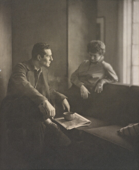 A black-and-white photograph of a White man and woman seated next to a sofa looking at one another.