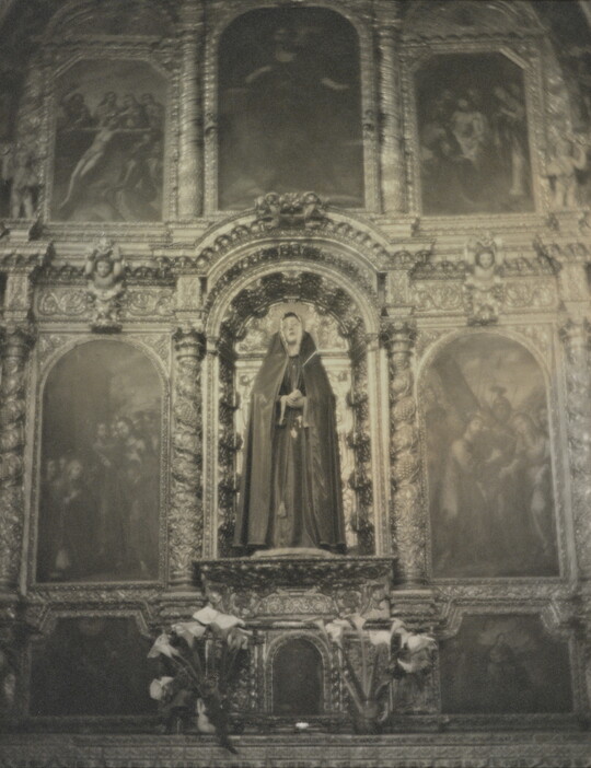 A black-and-white photograph of a sculpture of the Virgin Mary on an altar surrounded by paintings and decorative woodwork.