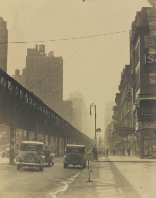 A sepia-toned photograph of vintage cars driving on a city street beside tall buildings and under an elevated train track.