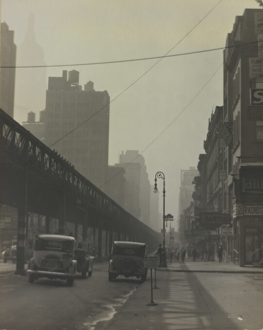 A black-and-white photograph of vintage cars driving on a city street beside tall buildings and under an elevated train track.