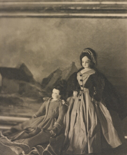 A black-and-white photograph of two light-colored dolls wearing formal 19th century European dresses posed in front of a landscape painting.