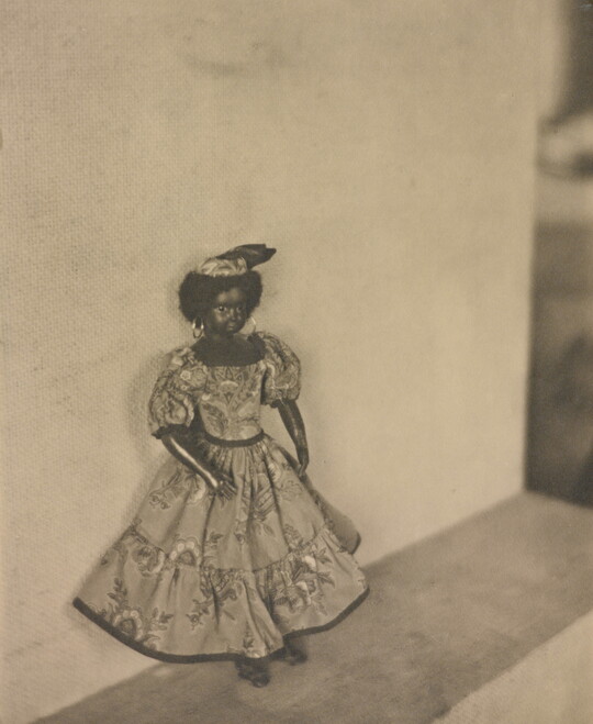 A black-and-white photograph of a dark-colored doll with curly hear wearing a patterned dress and hat posed against a wall.