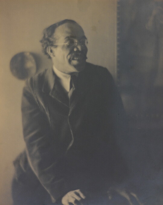 A sepia-toned, soft-focus photograph of a man wearing a suit and glasses leaning on something in front of him and looking away from the camera.