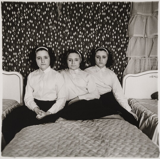 A black-and-white photograph of three teenage, triplet, girls dressed alike and seated on a twin bed.