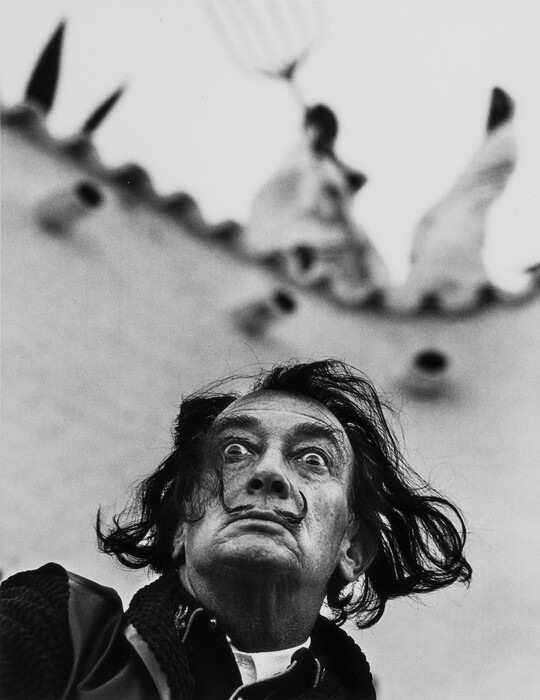 A black-and-white photograph looking up at a White man with medium-length wind-blown dark hair, a pencil mustache, wide eyes, and a frown.