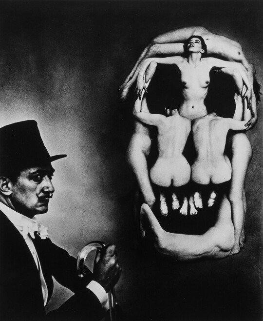 A black-and-white photograph of Salvador Dali in profile next to several nude women posed in the shape of a skull.