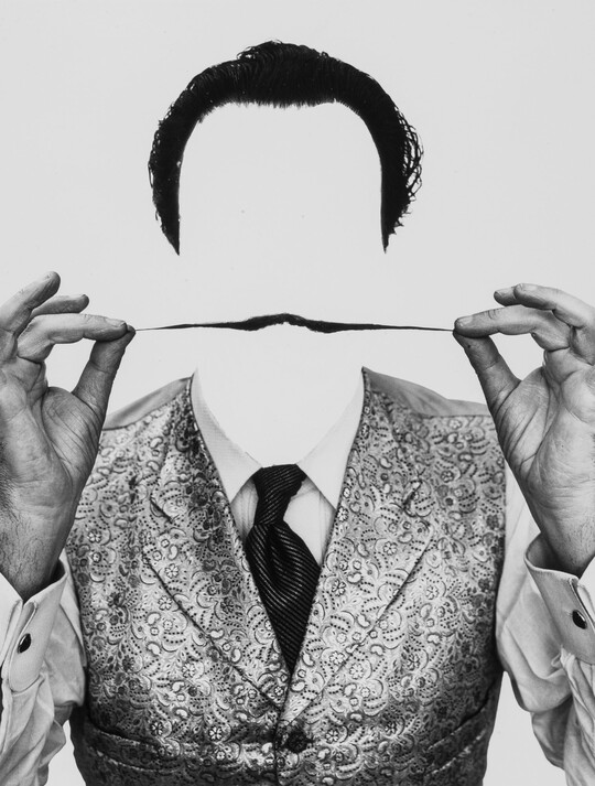 A black-and-white photograph of the head and shoulders of a man without a face holding out his pencil mustache with his hands.