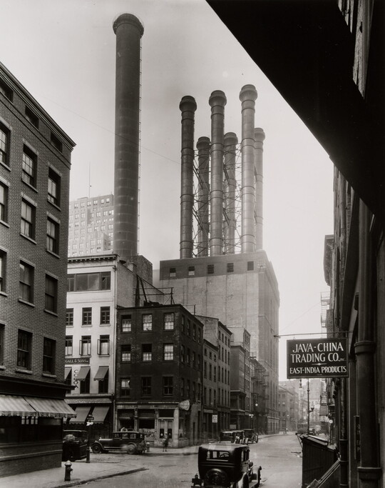A black-and-white photograph of a city street with buildings and factories with smokestacks.
