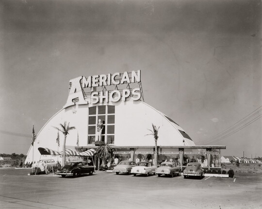 A black-and-white photograph of a building with a curved roof, a large sign that says "American Shops," and cars and fake palm trees in front of it.