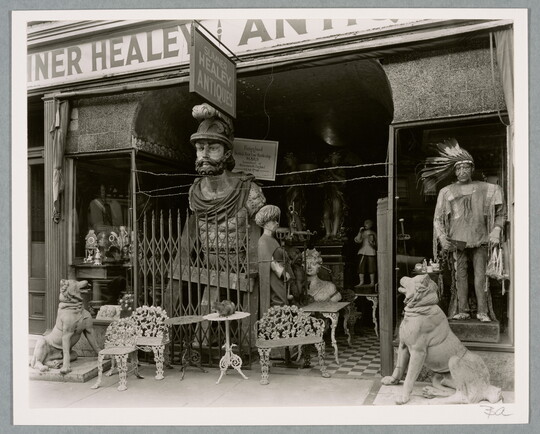 A black-and-white photograph of an overcrowded storefront with a variety of furniture and large statues at the entrance.