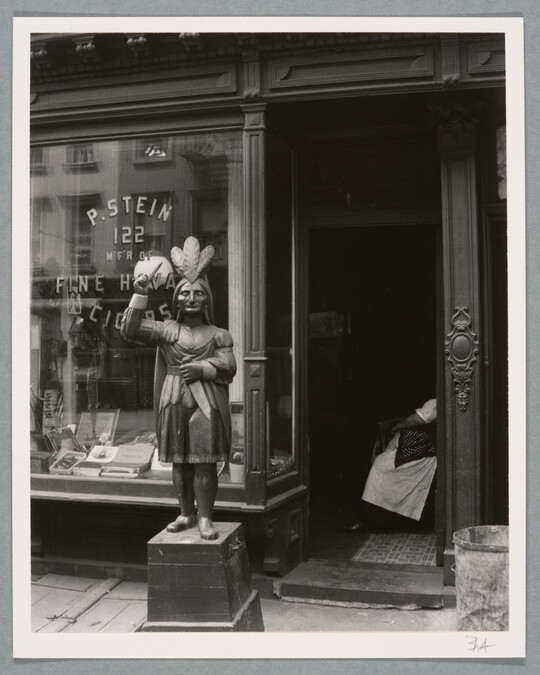 A black-and-white photograph of a storefront with a statue of an Indigenous person at the entrance.