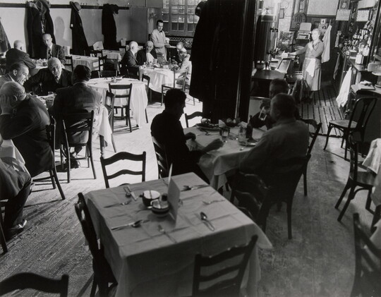 A black-and-white photograph of people sitting at multiple tables inside a restaurant.
