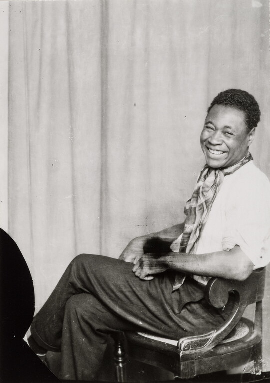 A black-and-white portrait of a Black man sitting with his legs crossed, smiling at the camera.